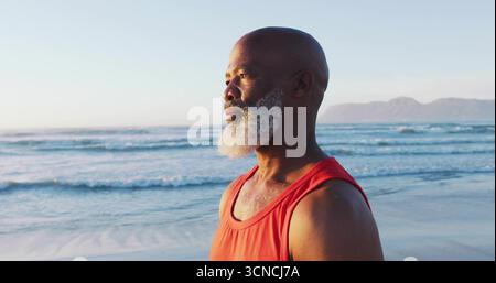 Standing senior man with white beard facing ocean at beach, with red sleeveless shirt, copy space Stock Photo