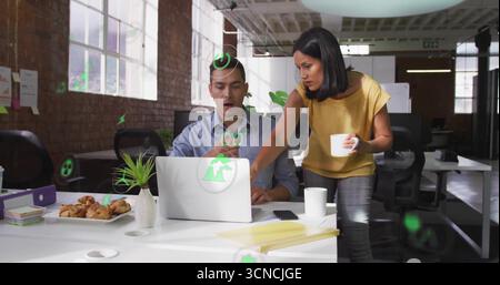 Collaborating pair reviewing green digital icons on laptop in modern loft office, with ceramic mugs Stock Photo