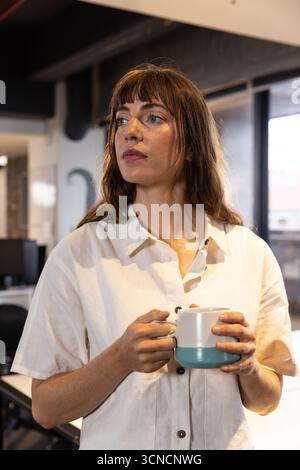 Mid adult woman holding two-tone ceramic mug gazing left in modern with monitors Stock Photo