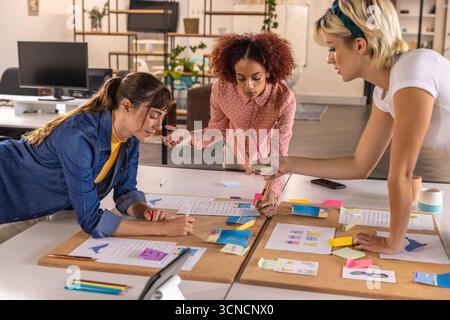 Diverse female coworkers collaborating on creative project at office with sticky notes, charts Stock Photo