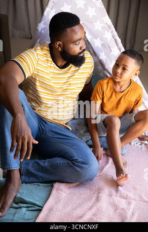 African American father and son sitting under star-patterned play tent on pastel cushions at home Stock Photo