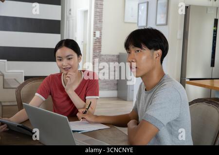 Asian friends studying together at dining table in home using laptop, tablet, printed papers Stock Photo