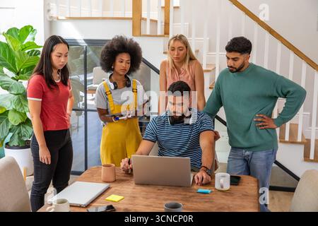 Diverse coworkers discussing project ideas around table in office using laptop and tablet Stock Photo