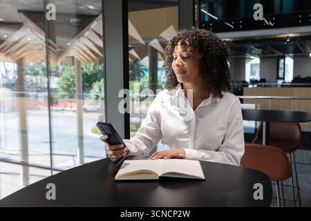 African american woman using smartphone, writing in notebook at lounge table in office Stock Photo
