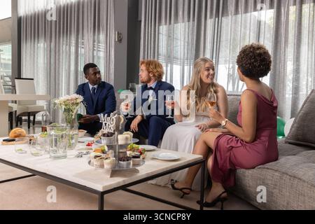Diverse newlywed couple with friends holding rosé glasses sitting on sofa at wedding reception Stock Photo
