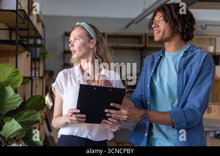 Diverse coworkers pointing at clipboard while discussing plan in shared office near potted plant Stock Photo