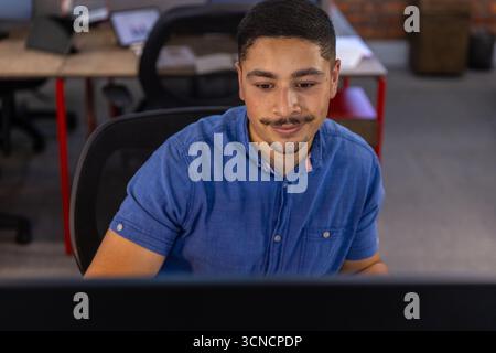 Man sitting on mesh chair at desk in modern open-plan office facing computer monitor, typing Stock Photo