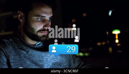 Bearded coder navigating user icons and blue notification bubble on laptop at home at night Stock Photo
