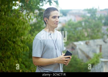 Millennial man using mobile phone with earbuds on viewpoint. Modern digital lifestyle concept showing adult enjoying wireless audio technology Stock Photo