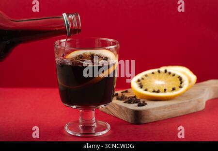 Pre-made mulled wine is poured from a bottle with a silver cap into a glass garnished with orange and cloves, evoking a cozy, accessible holiday feeli Stock Photo