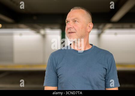 Portrait of a confident older man in a blue T-shirt, looking slightly upward and to the side with a calm, thoughtful expression. Stock Photo
