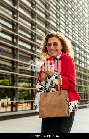Happy senior executive manager is holding a smartphone and smiling in front of an office building, enjoying a moment of relaxation during her workday Stock Photo