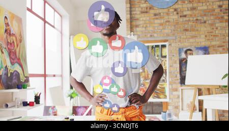 Standing mid adult Black male artist surveying art studio, with floating thumbs-up icons Stock Photo