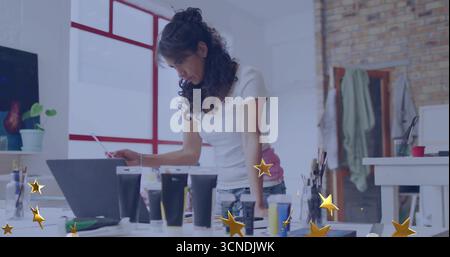 Leaning mature adult woman focusing on laptop screen in art studio, with paint tubes and brushes Stock Photo