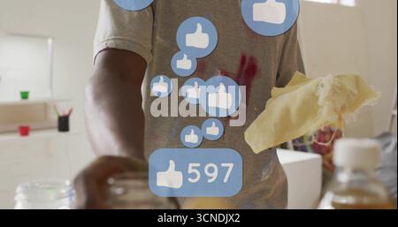 Wiping adult artist wearing paint-stained shirt in studio, with yellow sponge and thumbs-up icons Stock Photo