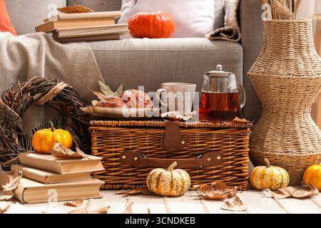 Autumn composition with teapot, cup of hot beverage, tasty buns and pumpkins on wicker basket in living room Stock Photo
