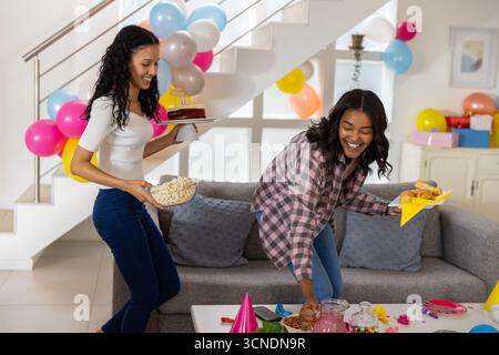 Diverse mom and teenage daughter carrying cake with candles and popcorn toward table Stock Photo
