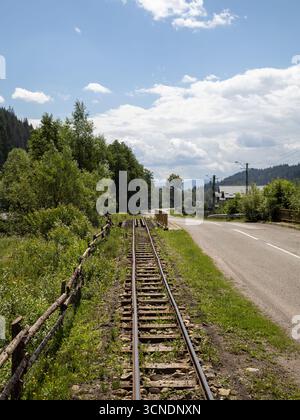 rustic scene showing an old, abandoned railway track running parallel to a road through a lush green landscape with mountains and forests under a blue Stock Photo