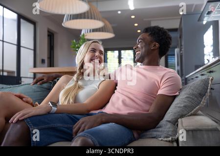 Diverse couple sitting on sofa under woven pendant lamps smiling in living room Stock Photo