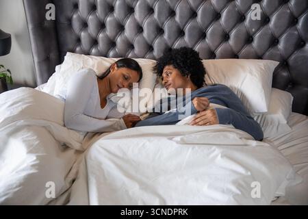 African american and asian indian couple lying under white duvet on bed, tufted headboard Stock Photo