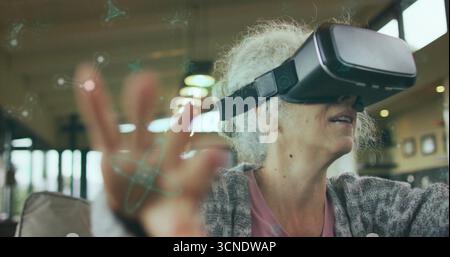 Interacting senior woman extending hand in modern lounge, with virtual reality headset, copy space Stock Photo