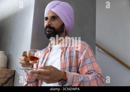 Indian man in lavender turban, standing in modern room next to staircase, holding glass tea cup Stock Photo