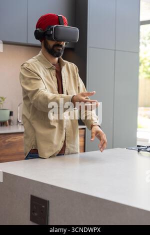Indian man using vr headset with headphones at kitchen island, manipulating virtual interface Stock Photo
