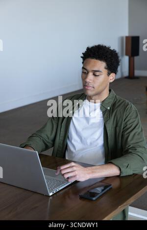 African american man working on silver laptop at table in study room using black smartphone Stock Photo