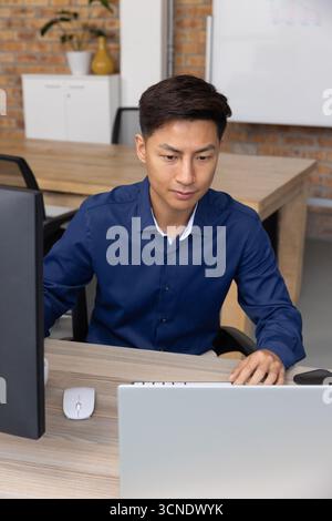 Asian male professional working on laptop and external monitor at office desk with mouse Stock Photo
