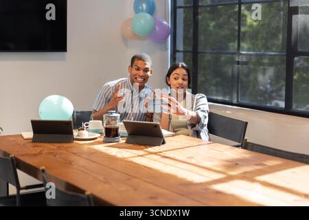 Diverse coworkers taking selfie with smartphone at meeting desk amid balloons, tablets, mugs Stock Photo