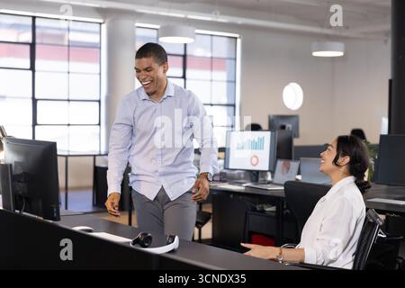 Diverse coworkers walking and sitting in open-plan office, female colleague smiling by monitors Stock Photo