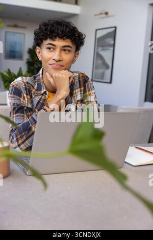 Asian man sitting at desk in home, using laptop, holding pen over notebook near plant Stock Photo