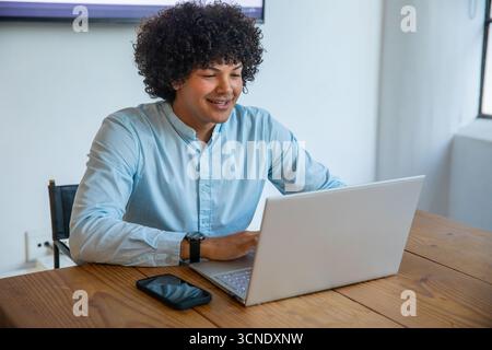 Man wearing light blue shirt sitting at home office desk typing on laptop near smartphone Stock Photo