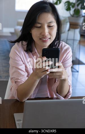 Asian woman sitting at wooden table holding smartphone while using laptop in living room Stock Photo