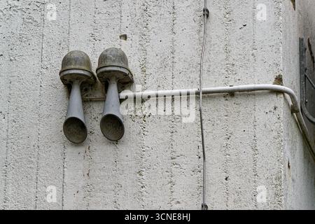 Two old loudspeakers are fixed high on a concrete wall. Wires run into a junction box and follow the surface toward the corner. Stains and rust show a Stock Photo