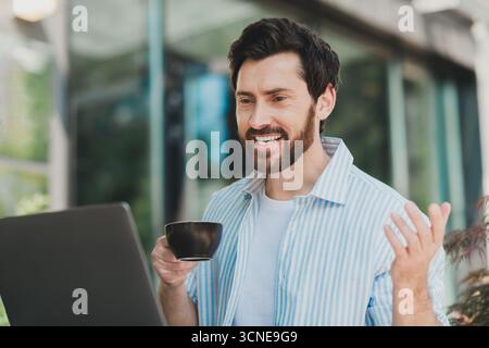 Young bearded man having an online conversation while working on laptop outdoors, holding a coffee cup, looking cheerful. Stock Photo