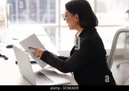 Happy middle aged accountant woman checking paper financial reports Stock Photo