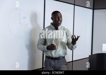 Male in boardroom pointing at pie chart on glass whiteboard with orange marker by lectern laptop Stock Photo