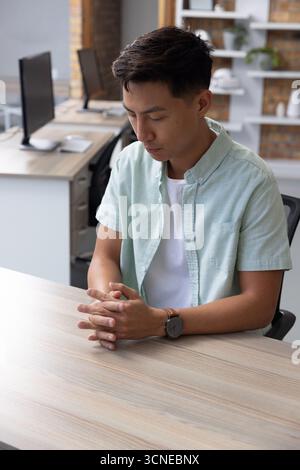 Asian man sitting at desk in modern open office, clasping hands and staring at notebook Stock Photo