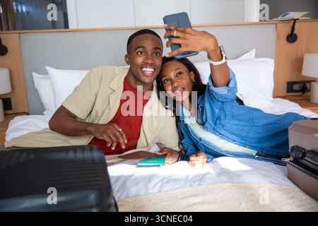 African American couple lying on hotel bed raising smartphone smiling with passports and suitcase Stock Photo