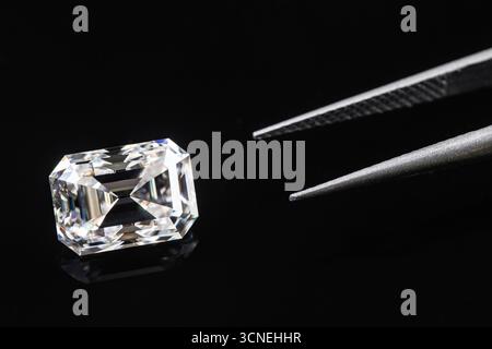 A diamond is on a table next to a pair of tweezers. The diamond is cut into a square shape Stock Photo