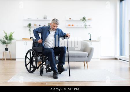 Disabled man practicing walking indoors using cane near wheelchair at home Stock Photo