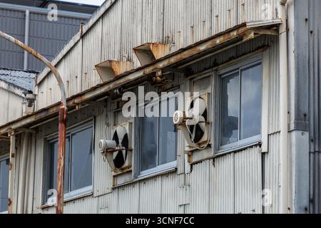Exterior ventilation fans mounted between windows sit silent on vintage factory's corrugated siding showing age with rust streaks and peeling paint. Stock Photo