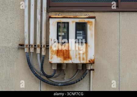 Weathered electrical box with visible rust affixed to beige wall, showing several insulated cables. Surface contrasts with simplicity of surroundings. Stock Photo