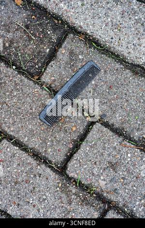 On pale paving tiles lies a small black comb, likely dropped or forgotten. Plastic teeth catch faint light, tiny scuffs show use. The quiet sidewalk s Stock Photo