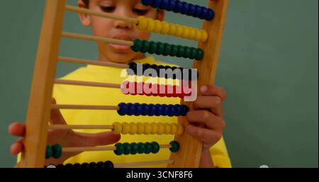 Boy wearing yellow shirt gripping abacus while focusing on colorful beads against green chalkboard Stock Photo