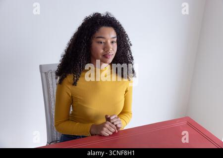 African american woman sitting at red table on chair and gazing off-camera in home workspace Stock Photo