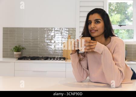 South Asian woman leaning on white kitchen island holding light brown ceramic mug, copy space Stock Photo
