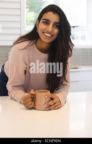 South Asian woman leaning on countertop in home kitchen while holding tan ceramic mug Stock Photo