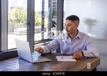 Man in late twenties working on laptop reviewing document at table in office, copy space Stock Photo